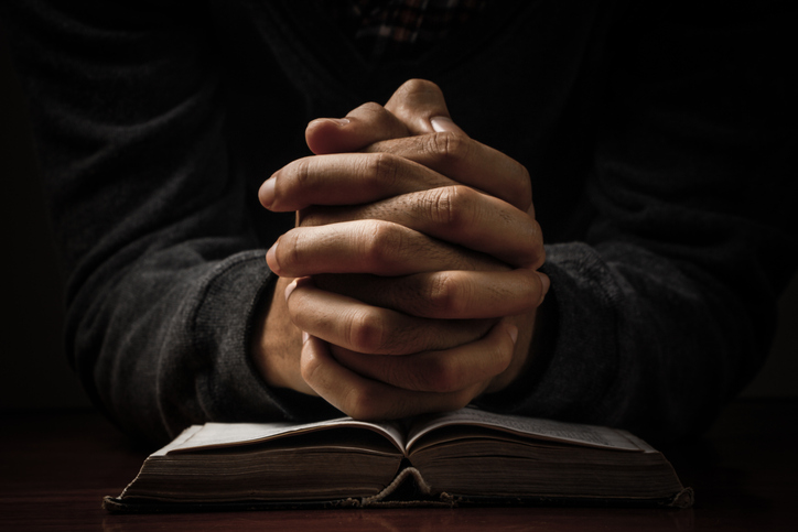 Hands clasped in prayer resting on an open Bible in dim lighting